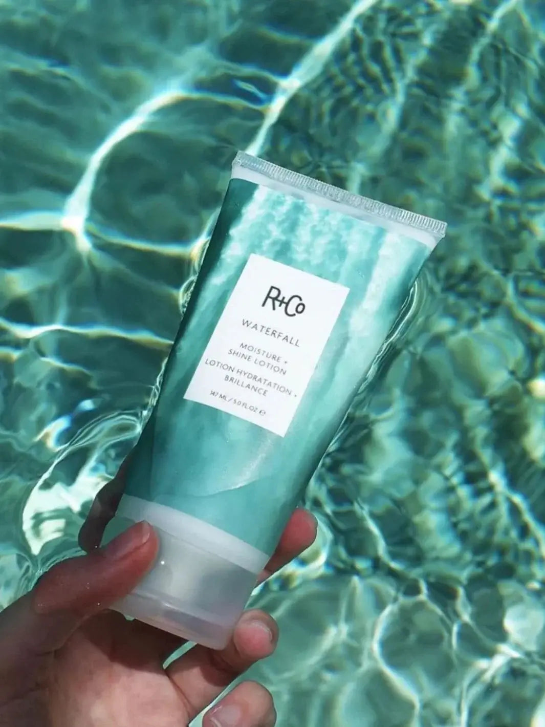 A hand holds a tube of R+Co WATERFALL Moisture + Shine Lotion directly above shimmering, sunlit pool water | Twentyseven Toronto