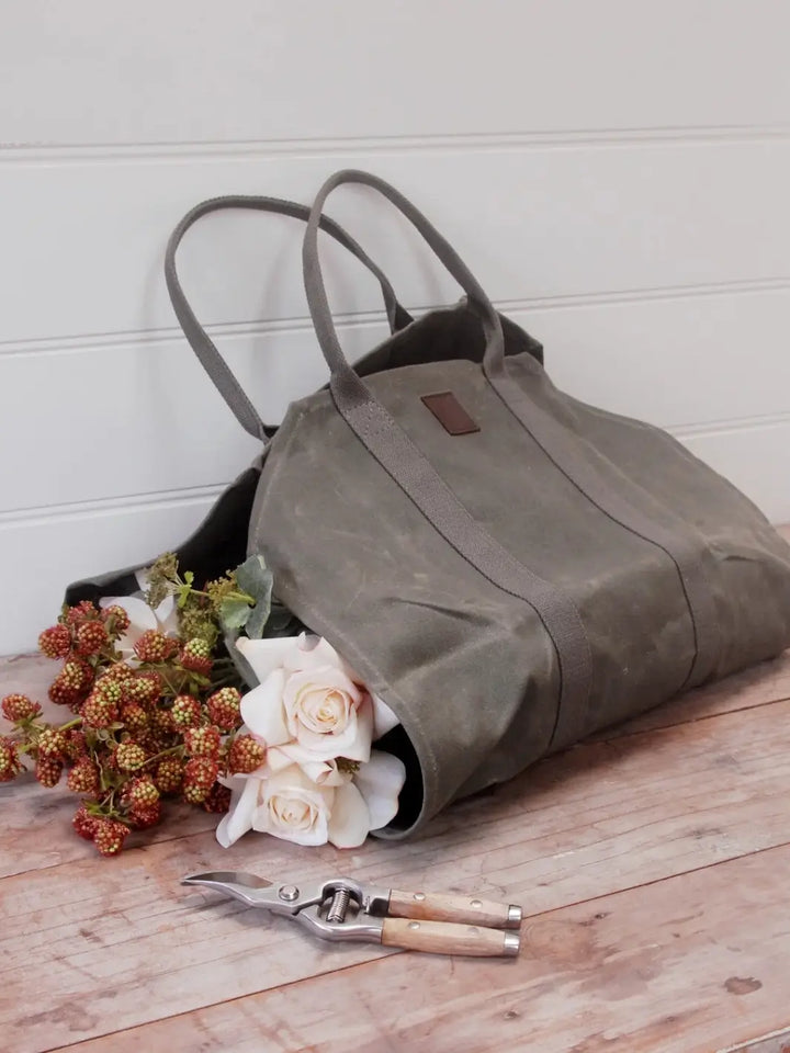 An olive green Waxed Canvas Log Flower Holder from Earth and Nest rests on a wooden surface next to garden shears, filled with white roses and textured botanical sprigs against a white wall | Twentyseven Toronto
