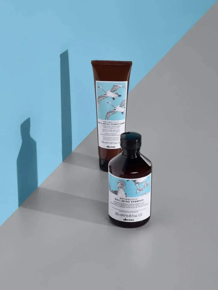 A bottle of Davines WELLBEING Shampoo stands on a grey surface in front of a matching tube of conditioner against a light blue backdrop | Twentyseven Toronto