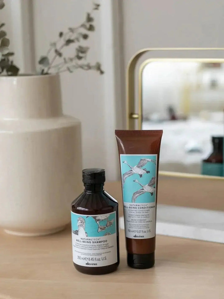 A bottle of Davines WELLBEING Shampoo and a tube of matching conditioner are placed on a wooden counter next to a ceramic vase | Twentyseven Toronto