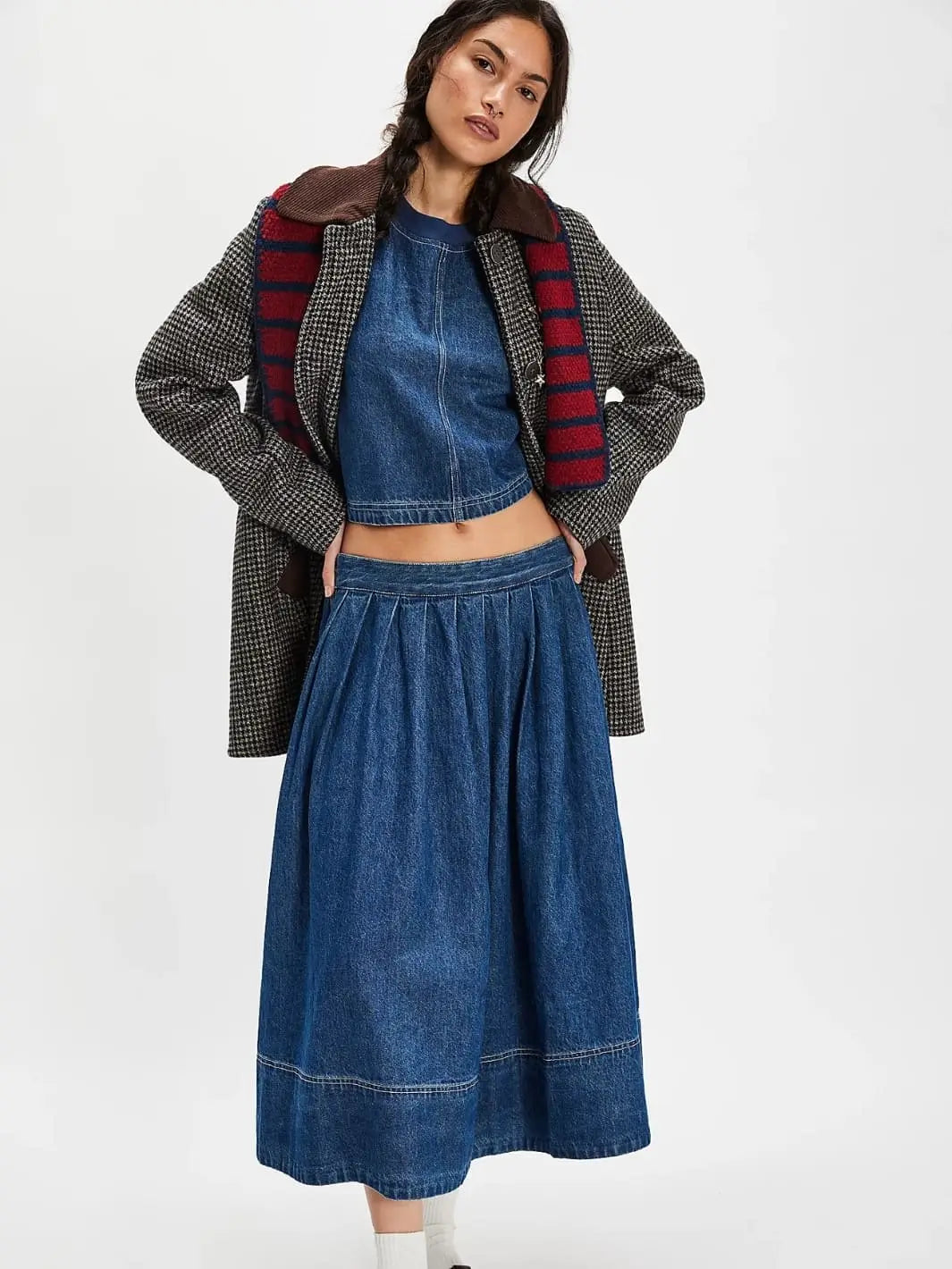 A full-length studio shot displays a model wearing the matching Free People Westley Denim Skirt Set in Deep Sea Wash styled under an unbuttoned checkered coat and a striped scarf | Twentyseven Toronto