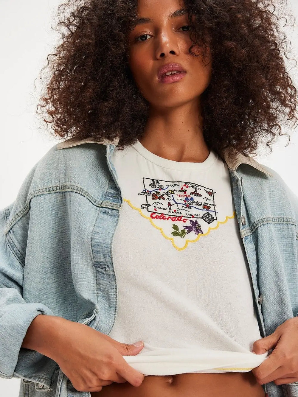 A model wears an oversized light-wash denim jacket layered over the slim-fitting Free People Wild Country Tee - Colorado Combo to showcase the embroidered front design | Twentyseven Toronto