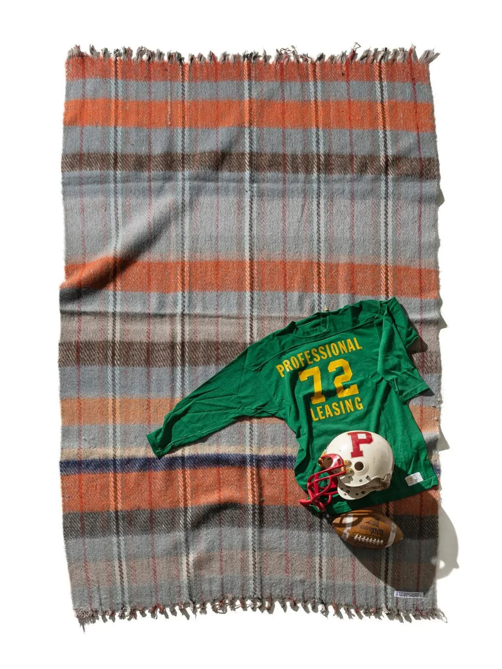 Puebco Recycled Wool Mix Blanket laid flat, featuring a grey, rust orange and brown plaid pattern with fringed edges, styled with vintage football gear | Twentyseven Toronto