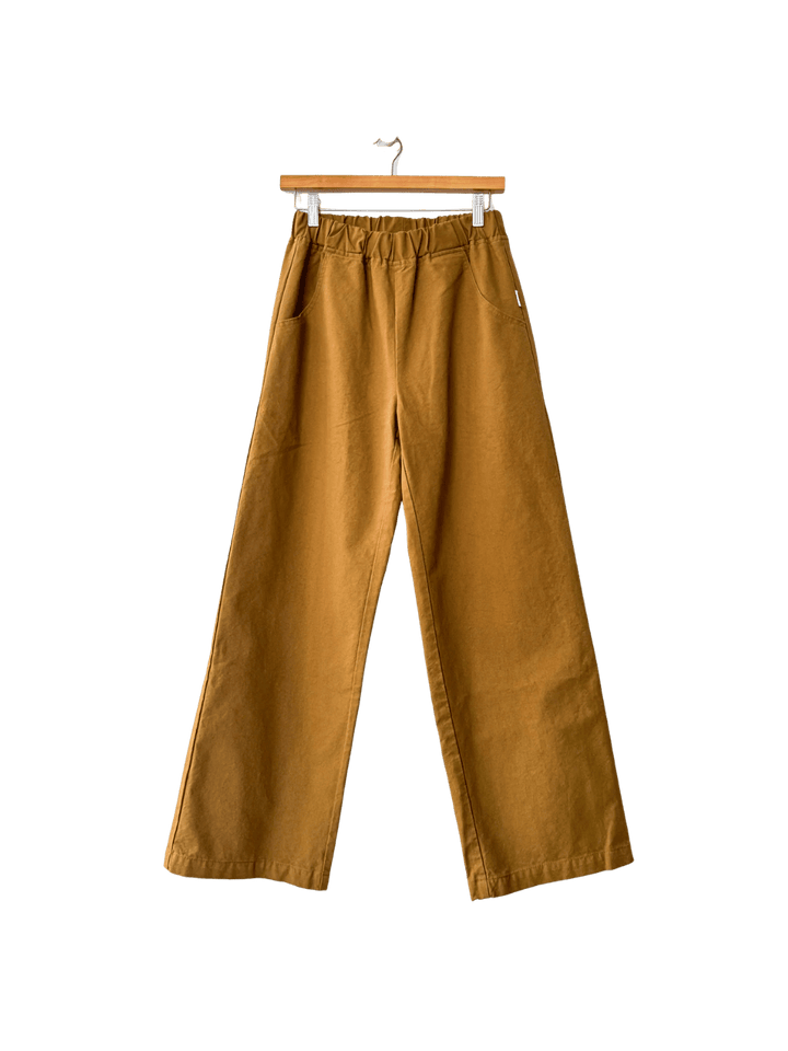 Mustard yellow Le Bon Shoppe 'Work Pants' in 'Dijon' hanging on a wooden hanger against a transparent background | Twentyseven Toronto