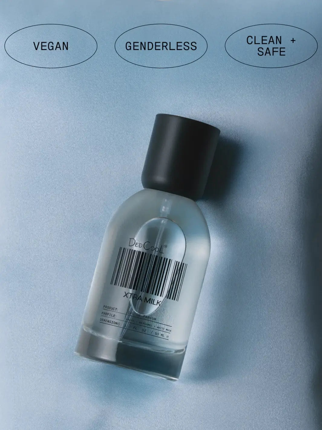 A 50ml full size bottle of Dedcool Xtra Milk Fragrance lying on a smooth blue fabric beneath circular labels reading "Vegan," "Genderless," and "Clean + Safe" | Twentyseven Toronto