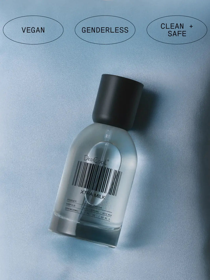A 50ml full size bottle of Dedcool Xtra Milk Fragrance lying on a smooth blue fabric beneath circular labels reading "Vegan," "Genderless," and "Clean + Safe" | Twentyseven Toronto