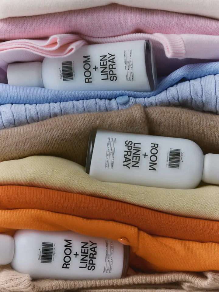 Three bottles including Dedcool Room + Linen Spray in Xtra Milk tucked horizontally between a colorful stack of folded sweaters and linens | Twentyseven Toronto