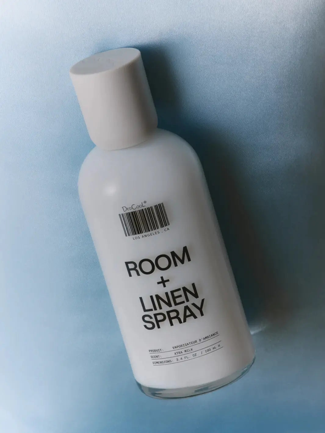 A bottle of Dedcool Room + Linen Spray in Xtra Milk resting diagonally against a soft, textured light blue background | Twentyseven Toronto