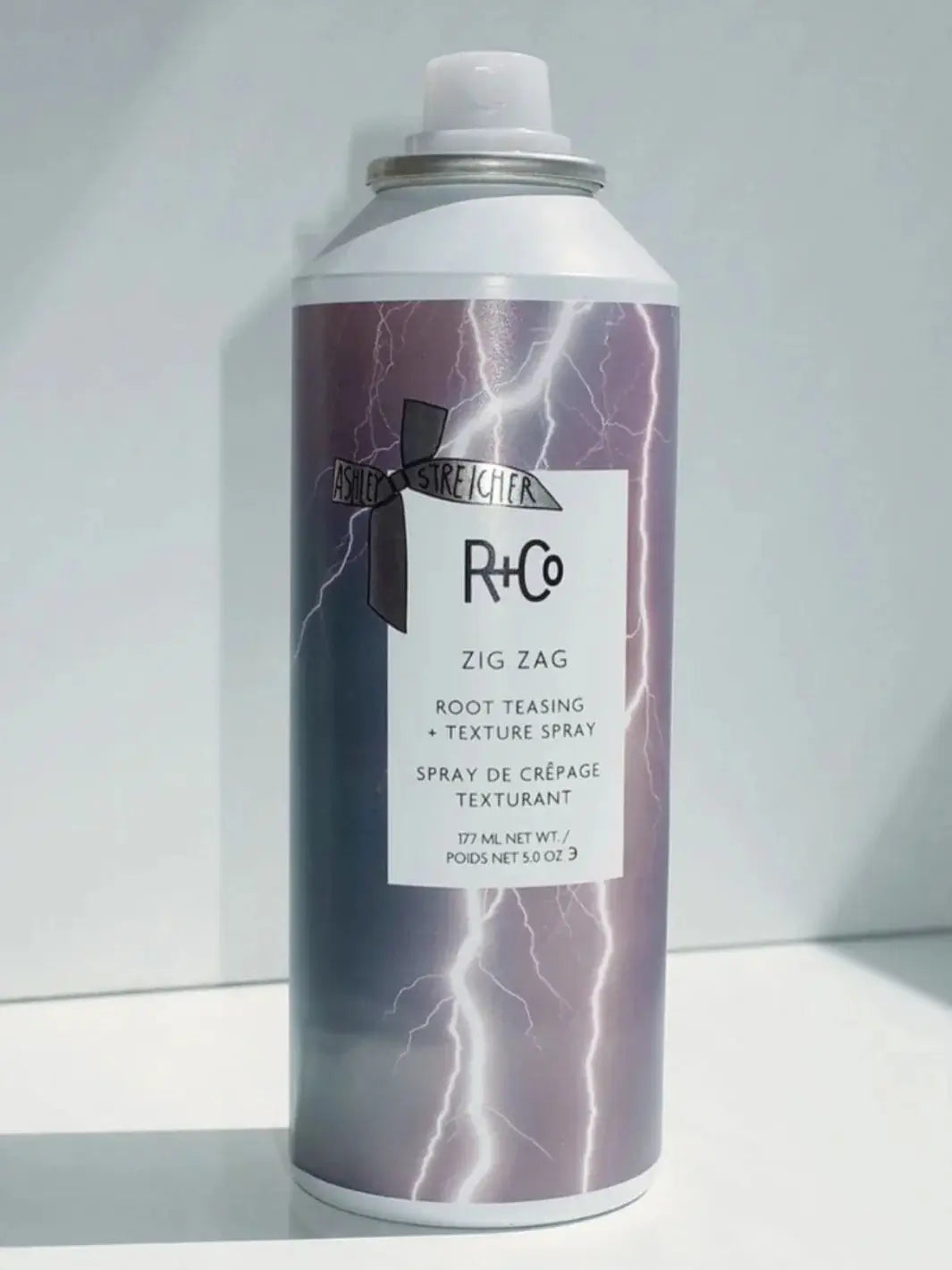 A can of R+Co ZIG ZAG Root Teasing and Texture Spray features a purple lightning graphic and stands upright against a white background | Twentyseven Toronto