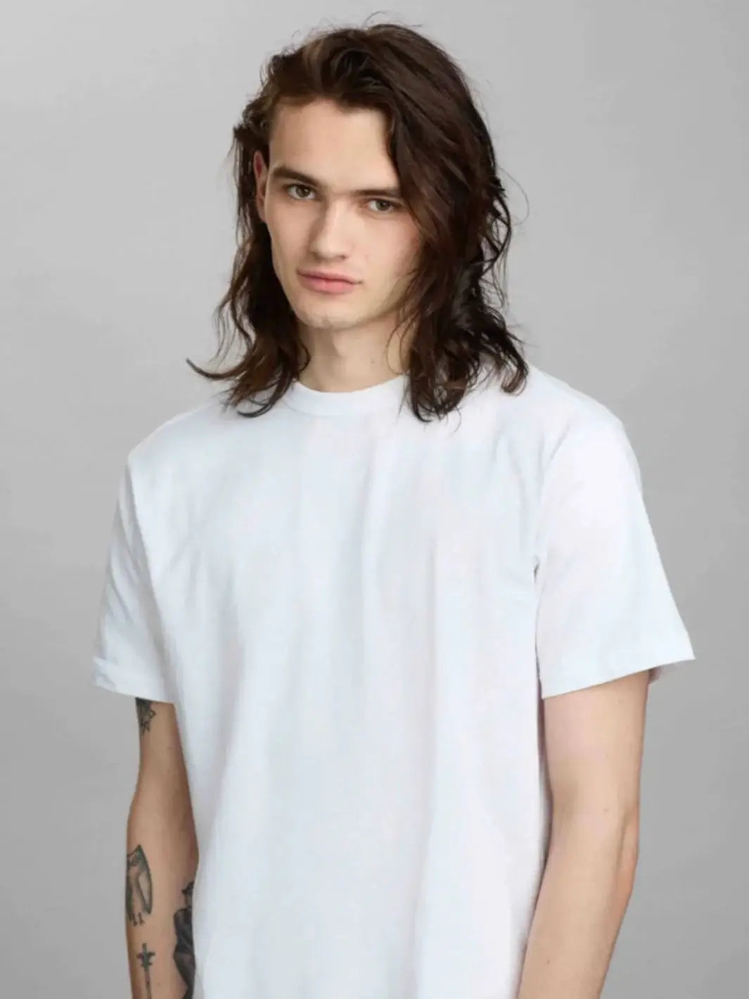 A model with shoulder-length, textured brown hair poses against a grey background, showcasing a look styled with R+Co ZIPPER Multitasking Styling Lotion | Twentyseven Toronto