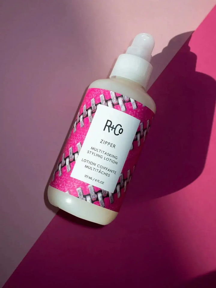 A bottle of R+Co ZIPPER Multitasking Styling Lotion with a bright pink zipper-print label lies diagonally across a dual-tone pink and magenta background | Twentyseven Toronto